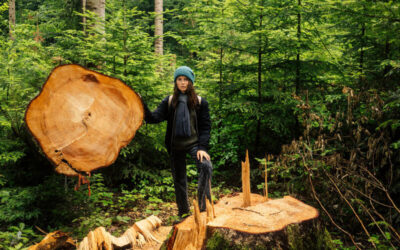 Una mujer con gorro de lana y ropa de abrigo posa junto a un árbol talado en medio de un bosque verde. Sostiene parte del tronco cortado, mostrando sus anillos de crecimiento, mientras se observa el tocón en primer plano.