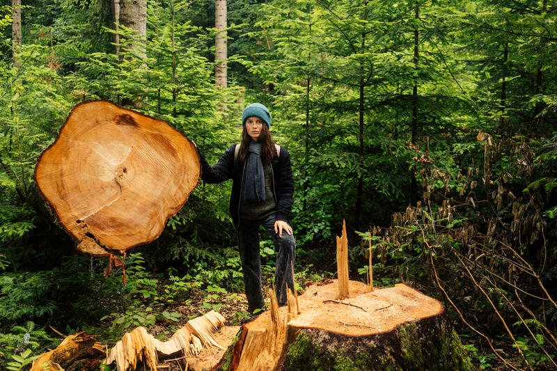 Una mujer con gorro de lana y ropa de abrigo posa junto a un árbol talado en medio de un bosque verde. Sostiene parte del tronco cortado, mostrando sus anillos de crecimiento, mientras se observa el tocón en primer plano.