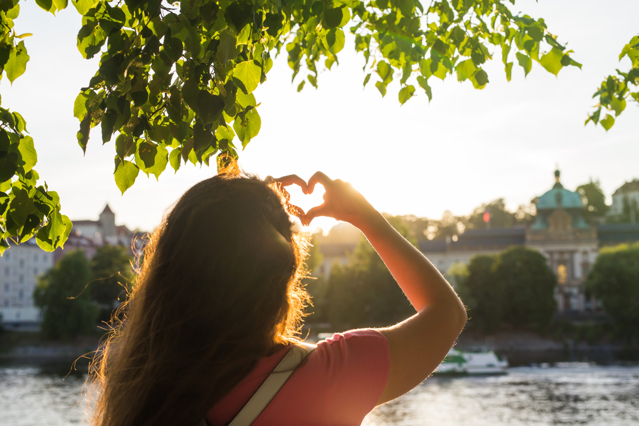 Mujer de espaldas bajo un árbol, haciendo un gesto de corazón con las manos hacia el sol, con un paisaje urbano y natural de fondo.