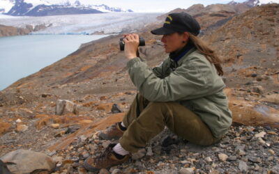 Guía de Parque Nacional observa con binoculares desde un terreno rocoso frente a un glaciar, en un entorno de montaña y lagos en la Patagonia.