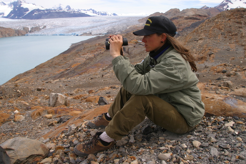 Guía de Parque Nacional observa con binoculares desde un terreno rocoso frente a un glaciar, en un entorno de montaña y lagos en la Patagonia.