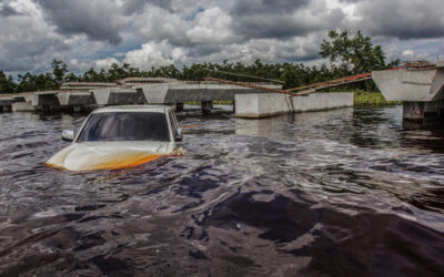 Una camioneta parcialmente sumergida en agua turbia frente a un puente colapsado, tras una grave inundación bajo un cielo nublado y amenazante.