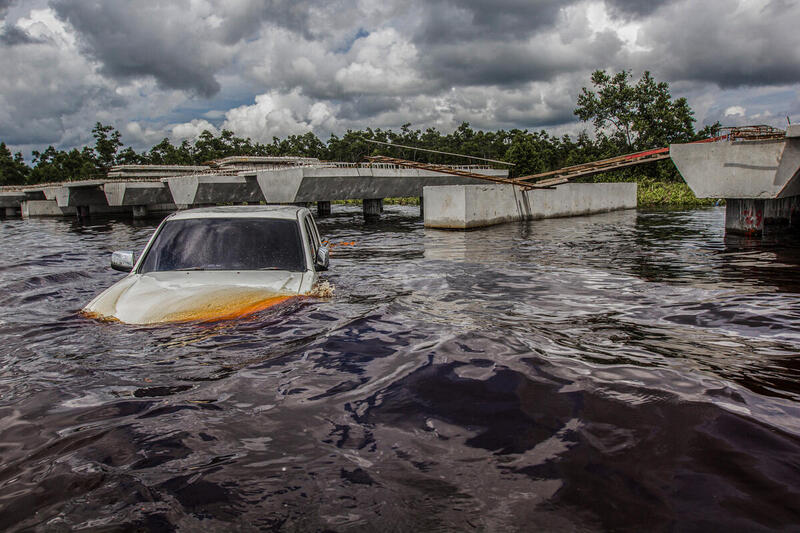 Una camioneta parcialmente sumergida en agua turbia frente a un puente colapsado, tras una grave inundación bajo un cielo nublado y amenazante.