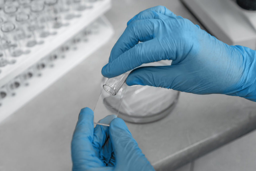 Primer plano de manos con guantes azules manipulando una lámina de vidrio y un tubo de ensayo en un entorno de laboratorio, representando procedimientos científicos.