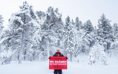 Persona en un bosque boreal nevado sostiene un cartel en idioma sámi, reclamando protección para los ecosistemas del norte.