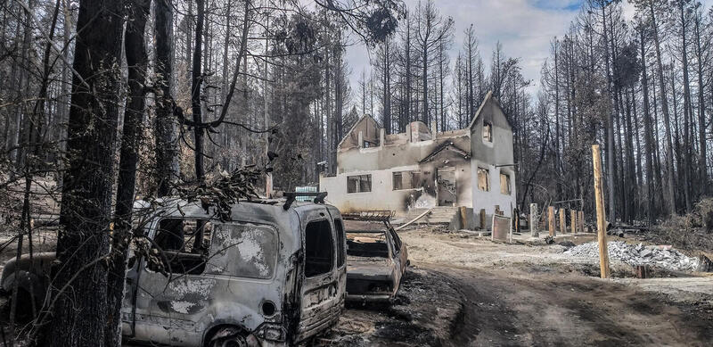 Viviendas y vehículos destruidos por un incendio forestal en una zona boscosa de la Patagonia argentina, con árboles calcinados y humo en el ambiente.