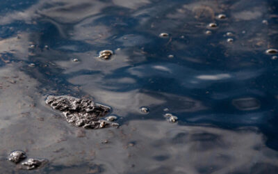 Mancha oscura de hidrocarburos flotando sobre una superficie de agua contaminada, con burbujas y residuos visibles.