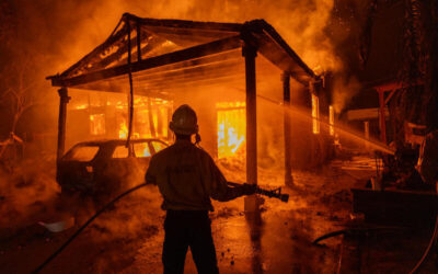 Bombero con manguera intentando apagar un incendio que consume una casa y un automóvil.