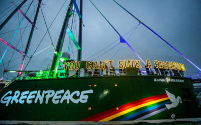Barco Rainbow Warrior de Greenpeace iluminado por luces de colores en el puerto, con la frase “You can’t sink a rainbow” sobre la cubierta.