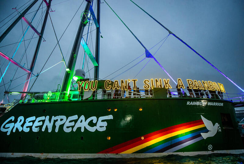 Barco Rainbow Warrior de Greenpeace iluminado por luces de colores en el puerto, con la frase “You can’t sink a rainbow” sobre la cubierta.