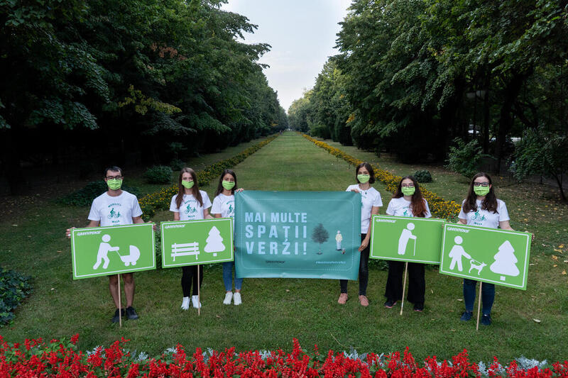 Grupo de jóvenes con mascarillas verdes sostiene carteles que promueven la creación de más áreas verdes en una ciudad, en medio de un parque arbolado.